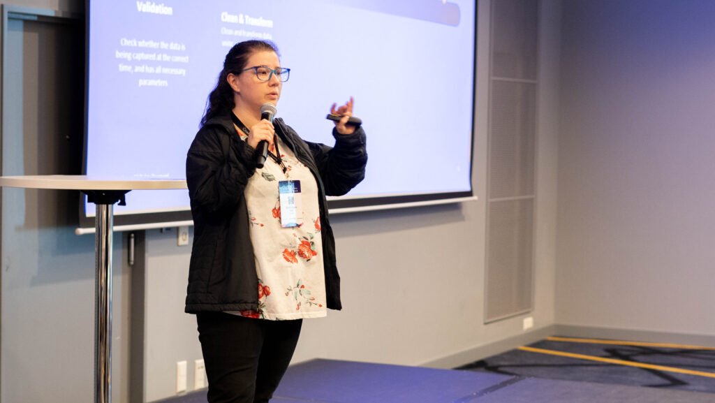 A speaker presenting with slides behind her